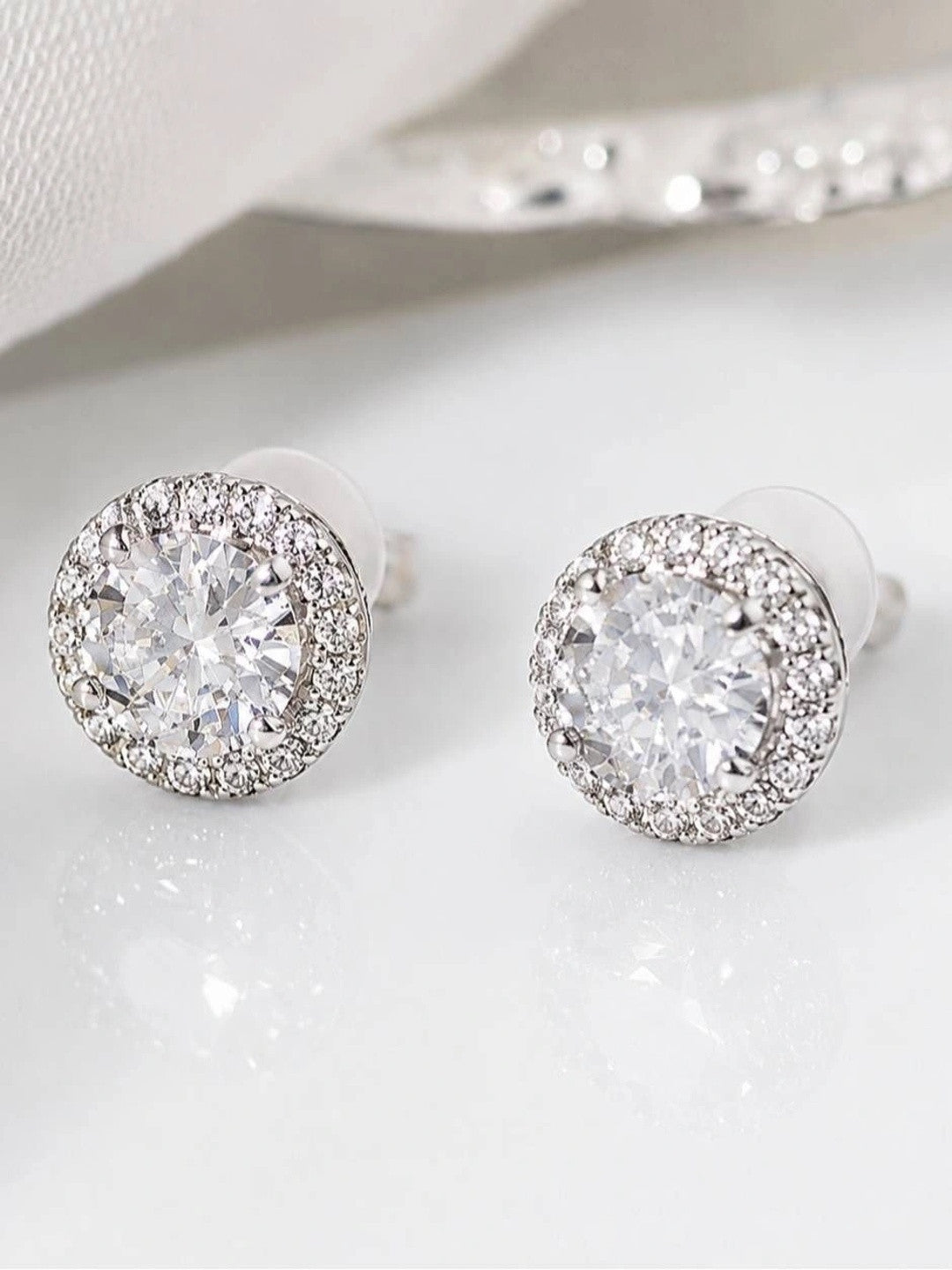 Pair of silver-finish CZ halo stud earrings with round solitaire centre and pavé surround, on white surface