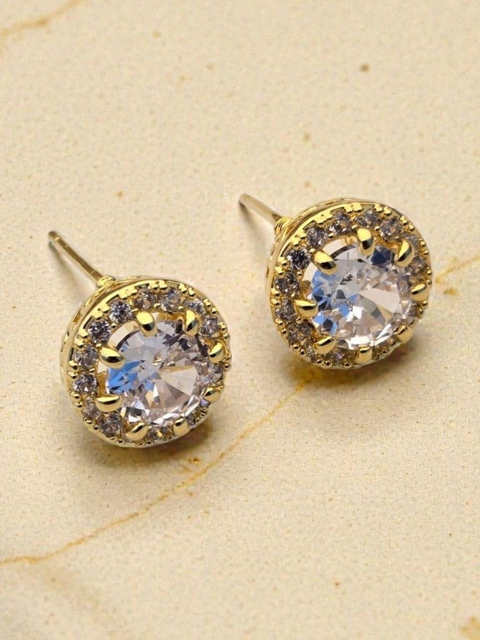 Close-up front view of gold-plated halo stud earrings with round CZ centre on beige surface