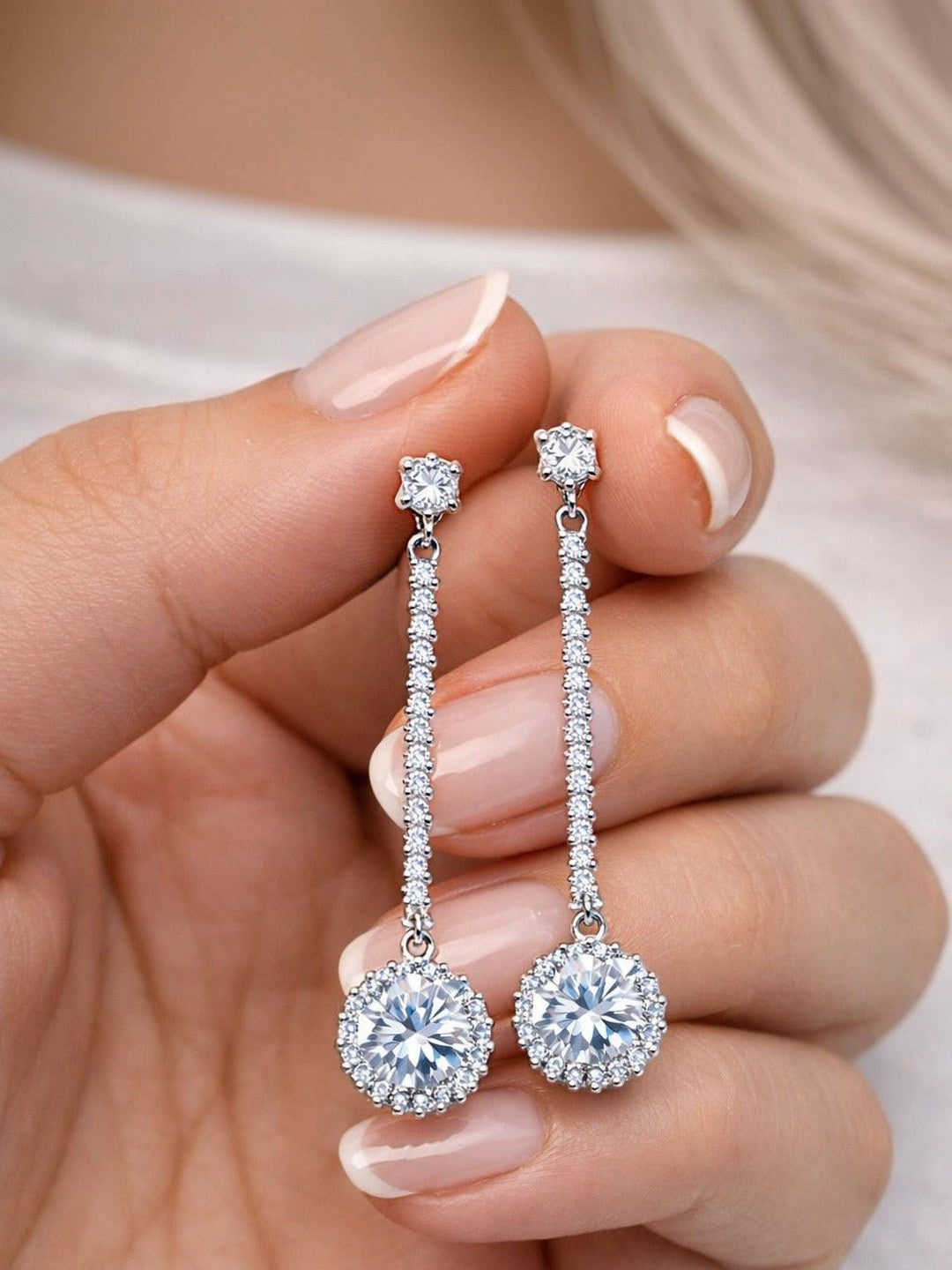 Hand holding a pair of silver CZ long drop earrings with halo pendant and pavé bar against a soft blurred background