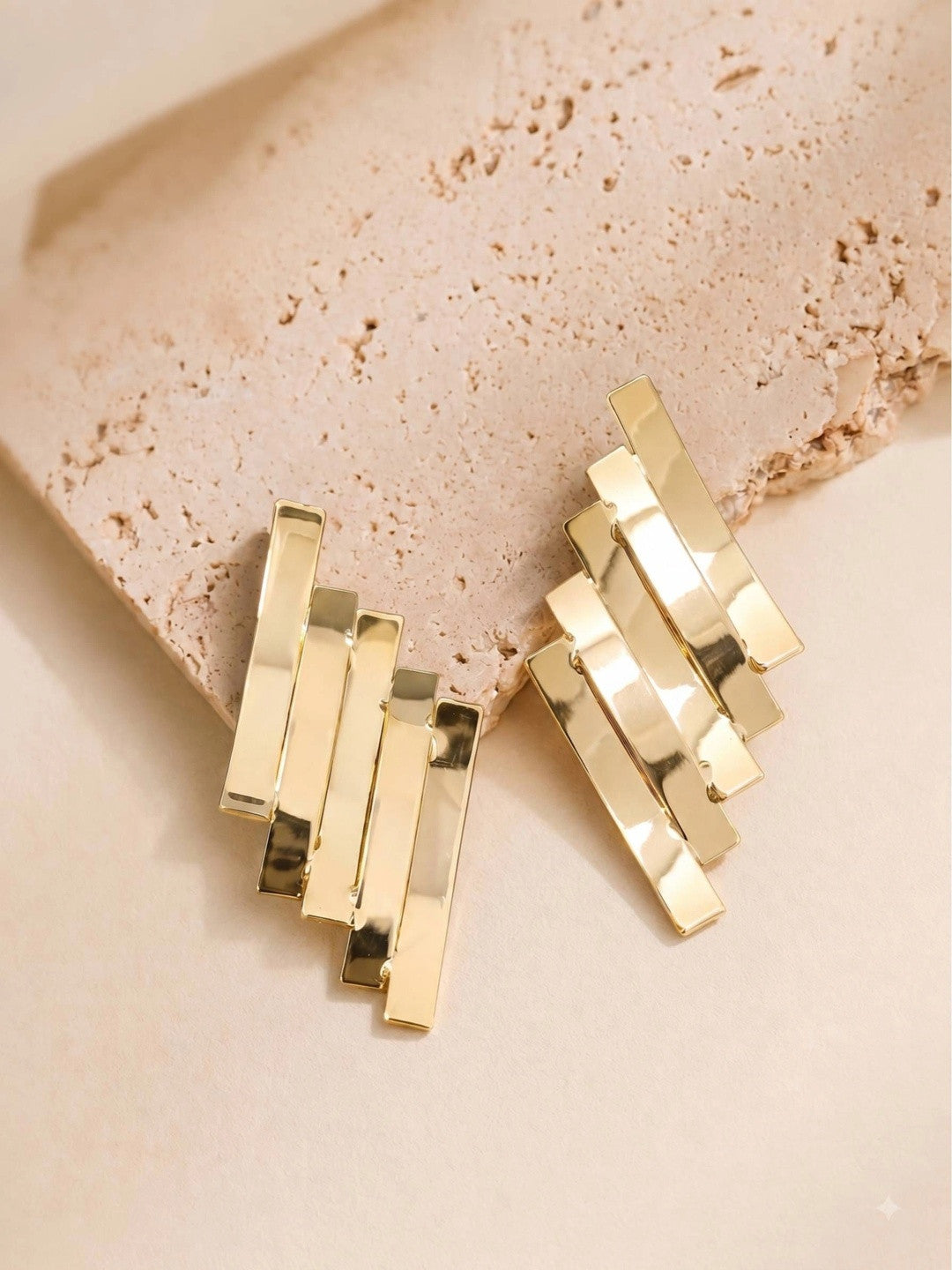 Gold geometric layered bar earrings resting on a textured beige stone surface