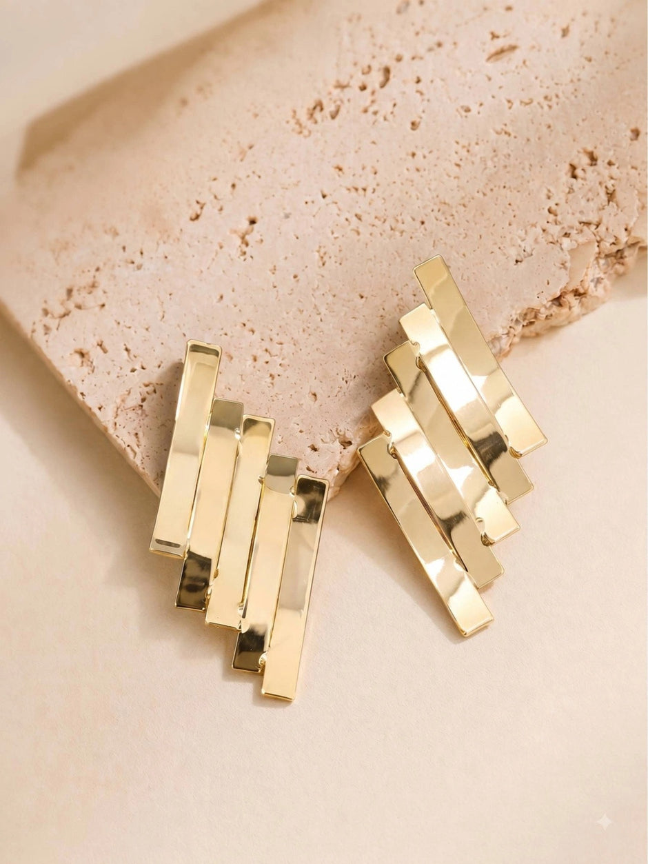 Gold geometric layered bar earrings resting on a textured beige stone surface