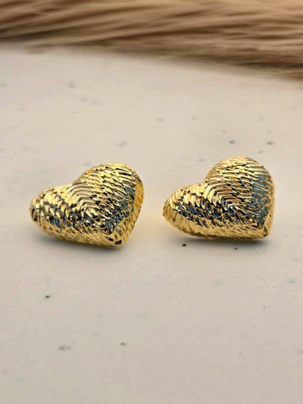 Pair of gold-plated ridged heart stud earrings on a white marble surface with dried grass in background