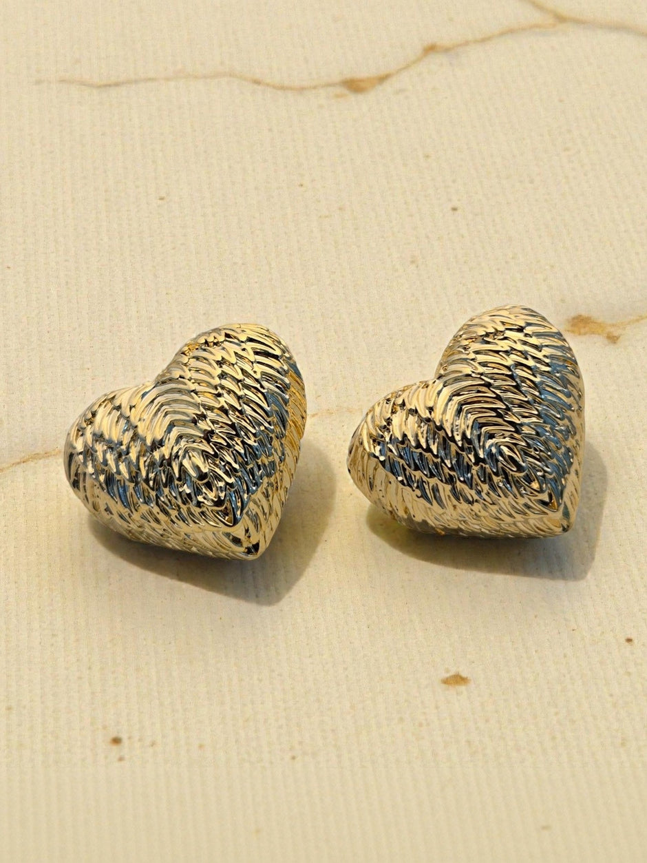 Close-up of gold-plated diagonal-ridged heart studs placed on a beige linen surface