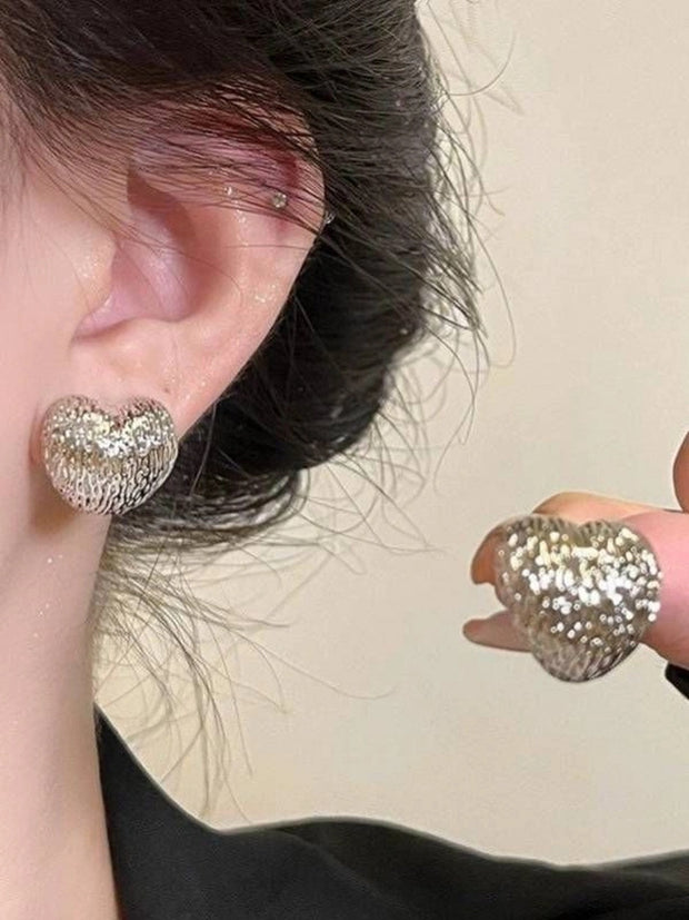 Silver textured heart stud worn on a model's ear with second earring held between fingers