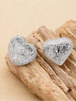 Silver ridged heart stud earrings displayed on driftwood against a sandy beige background