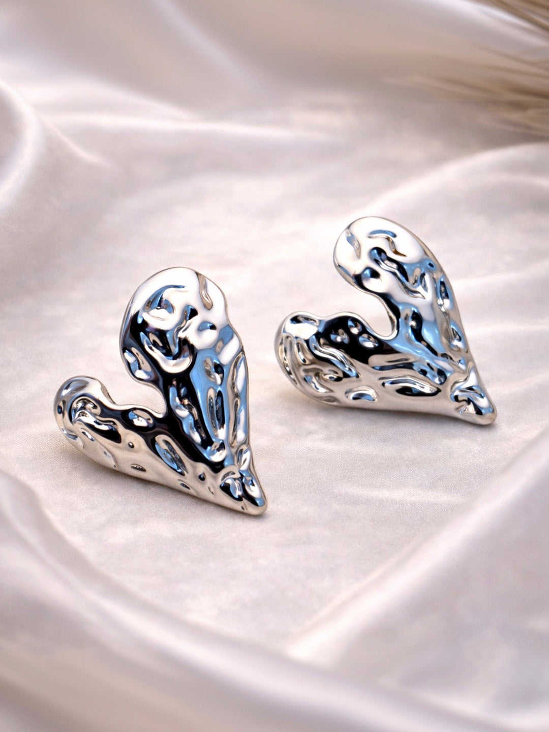 Pair of silver-finish molten heart stud earrings resting on soft white satin fabric