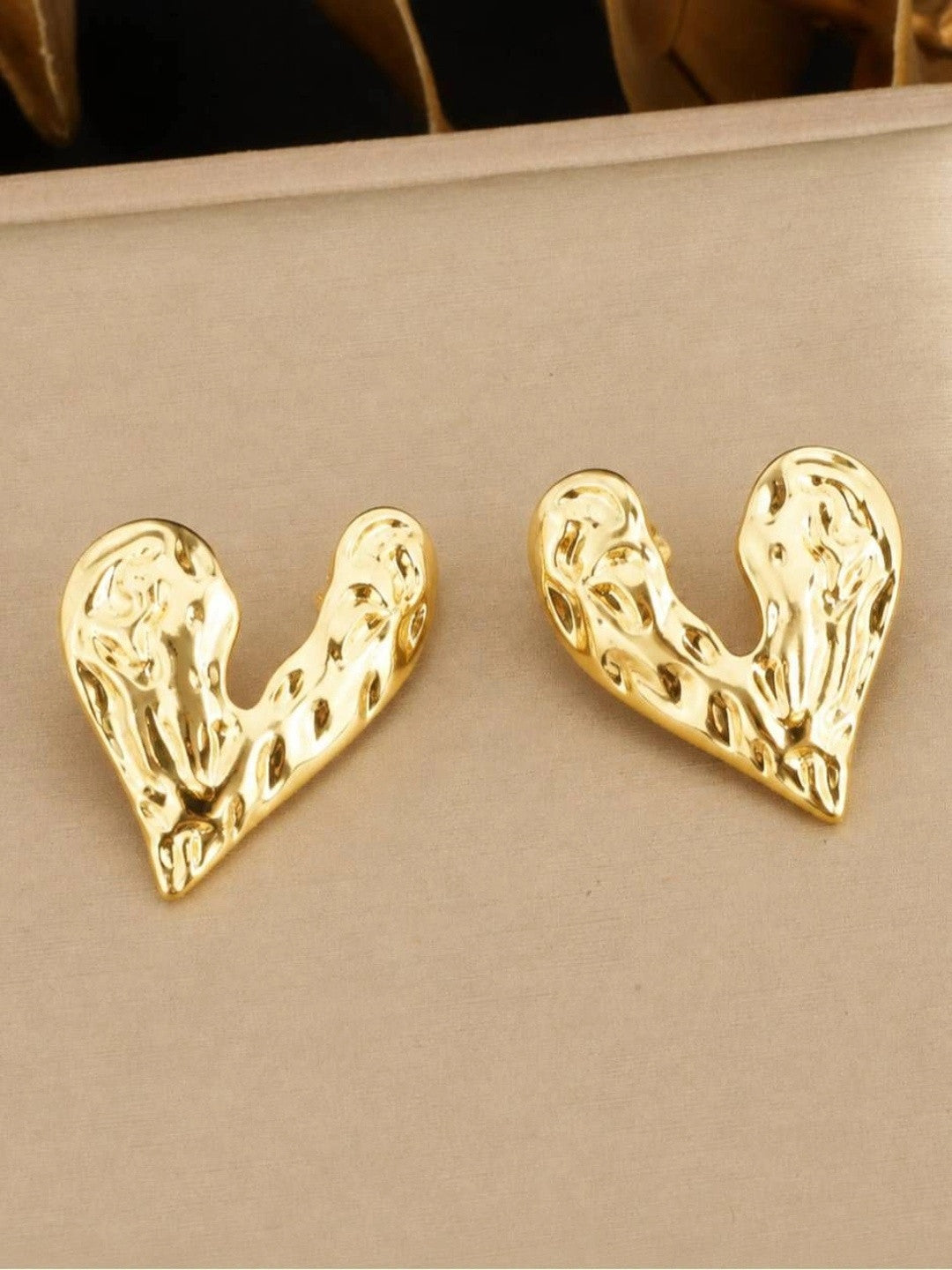 Pair of gold-plated hammered heart stud earrings laid flat on a beige surface