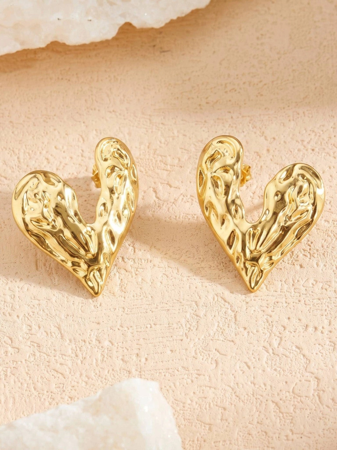 Both gold hammered heart stud earrings displayed upright on a textured pink stone surface