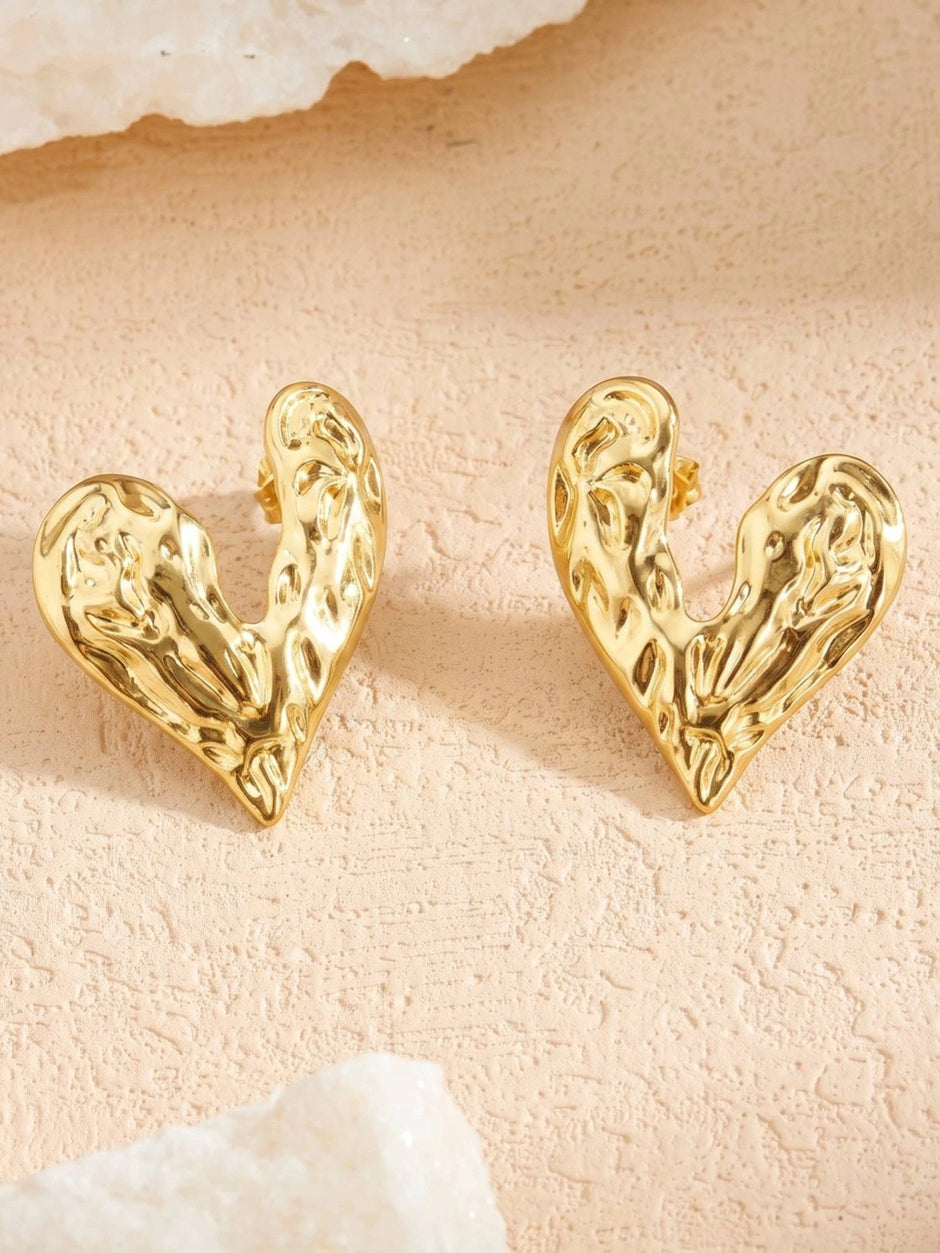 Both gold hammered heart stud earrings displayed upright on a textured pink stone surface