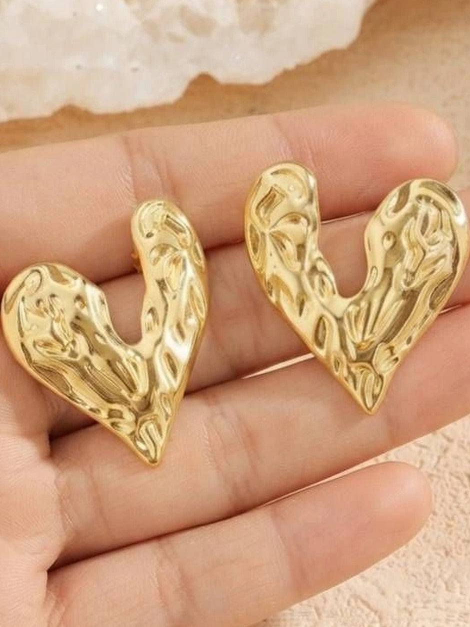 Two gold hammered heart stud earrings resting in a model's open palm for size reference