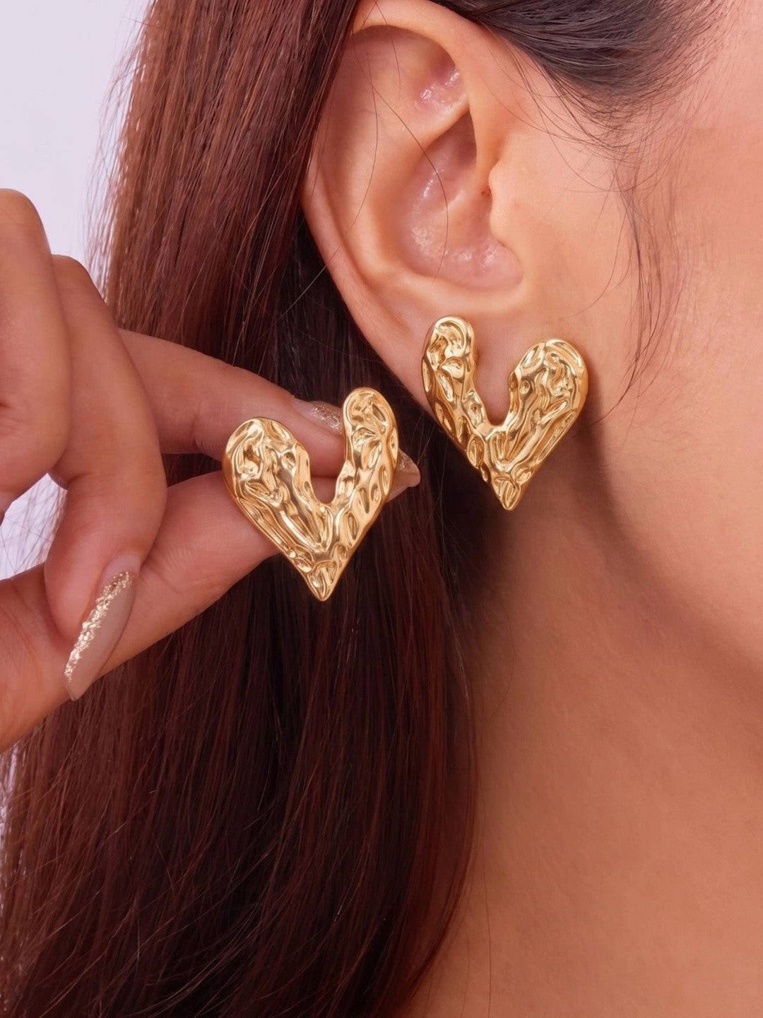 Model wearing oversized gold hammered heart stud earrings on both ears, hair tucked back