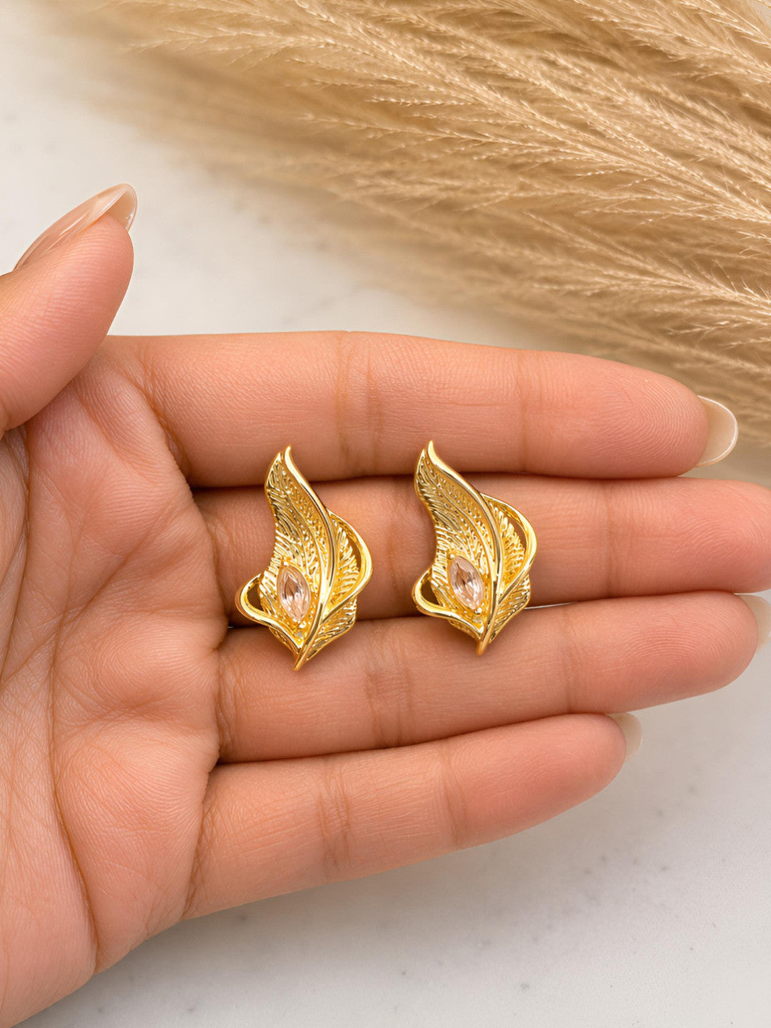 Two gold feather stud earrings with blush marquise crystals resting on an open palm