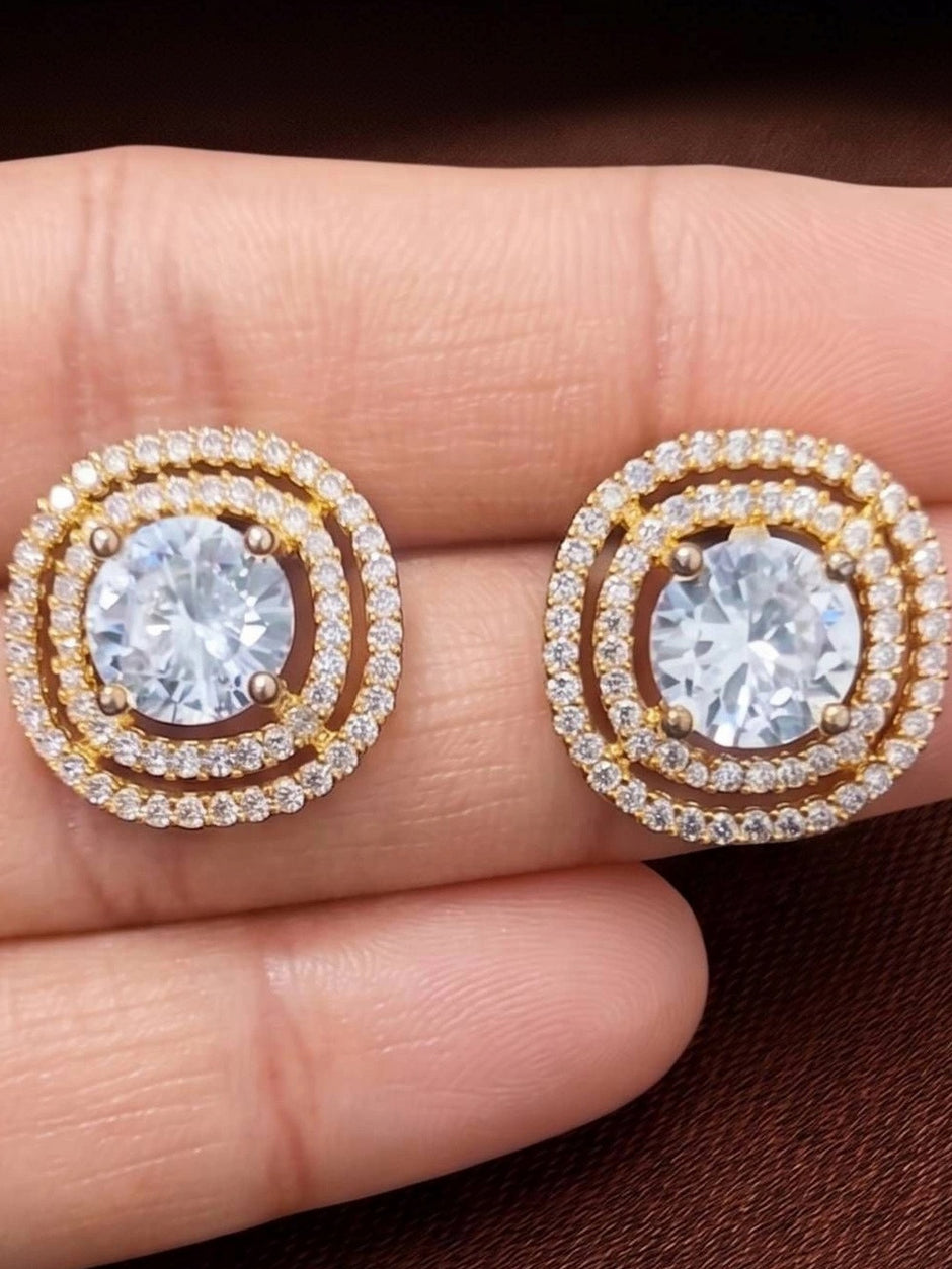 Close-up of two gold-plated double halo CZ stud earrings held between finger and thumb