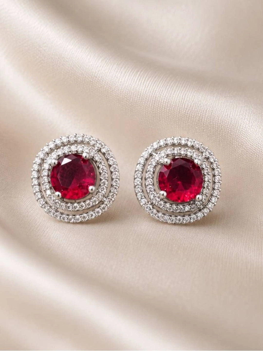 Pair of silver-tone double halo ruby red stud earrings laid flat on cream satin fabric