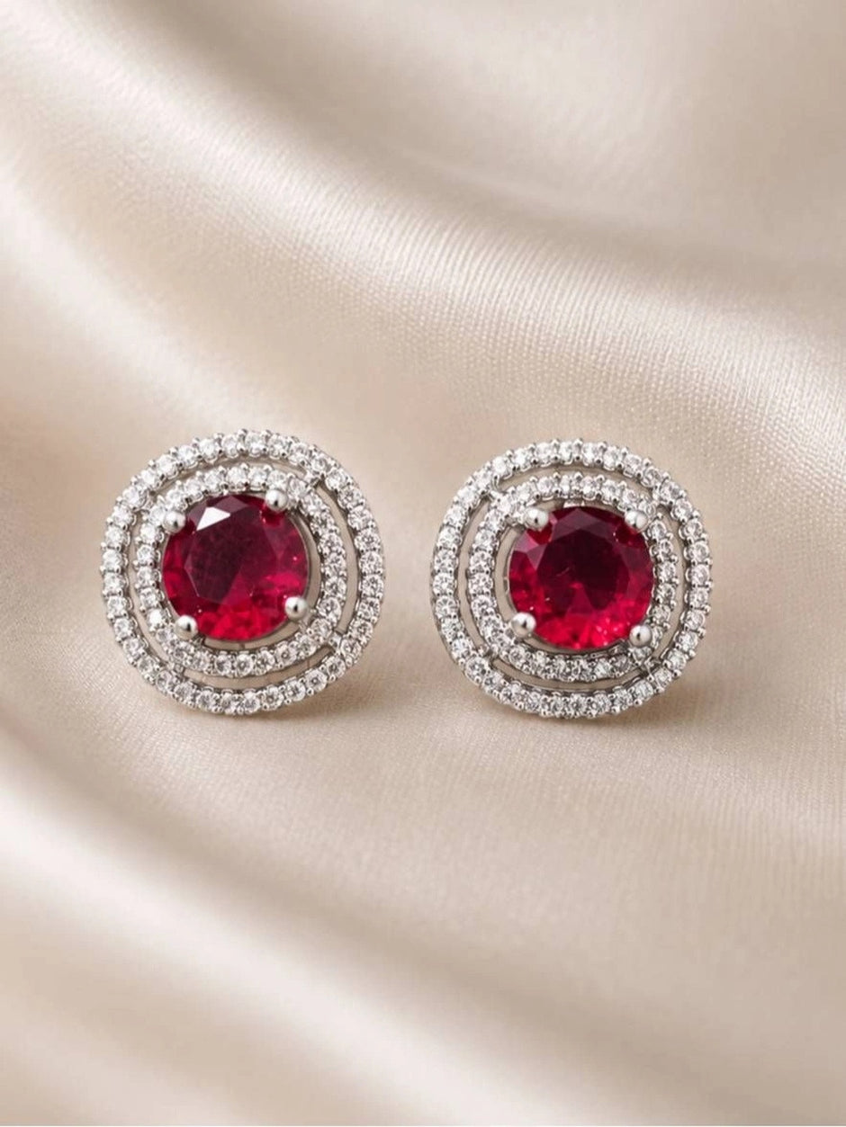 Pair of silver-tone double halo ruby red stud earrings laid flat on cream satin fabric