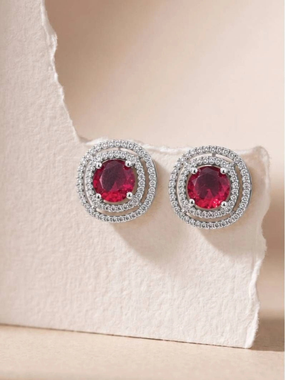 Two ruby red double halo stud earrings resting on a white textured stone surface