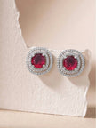 Two ruby red double halo stud earrings resting on a white textured stone surface