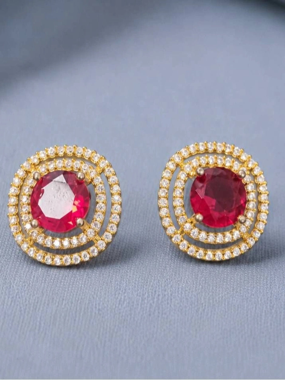 Pair of gold-plated double halo stud earrings with round ruby-red CZ centre stones on grey fabric