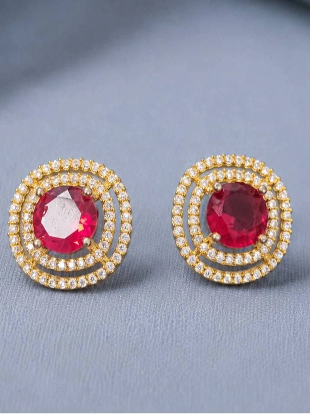 Pair of gold-plated double halo stud earrings with round ruby-red CZ centre stones on grey fabric