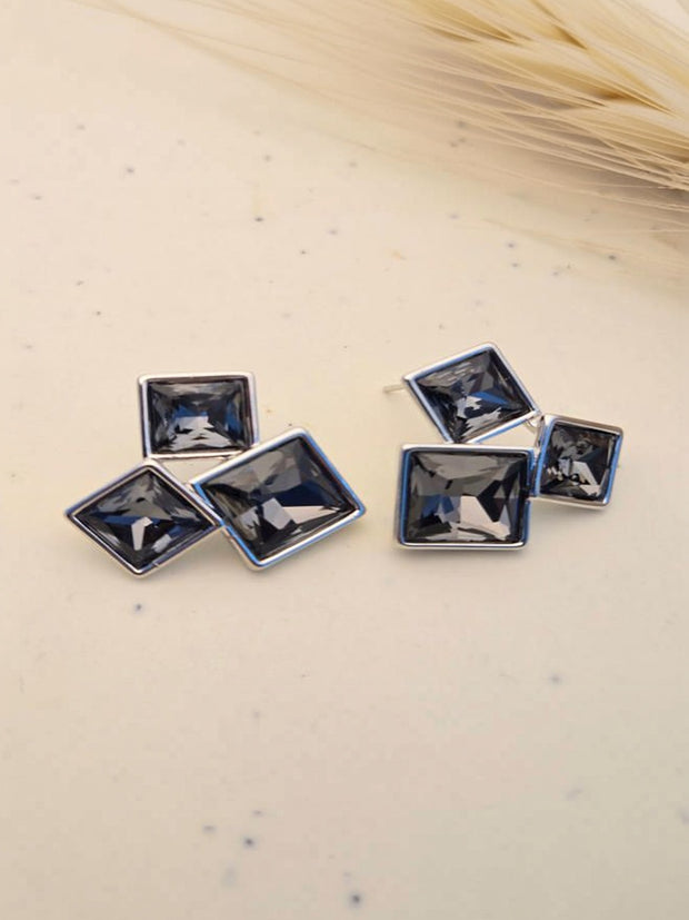 Pair of silver-tone blue crystal square cluster stud earrings laid flat on a cream speckled surface with dried grass in background