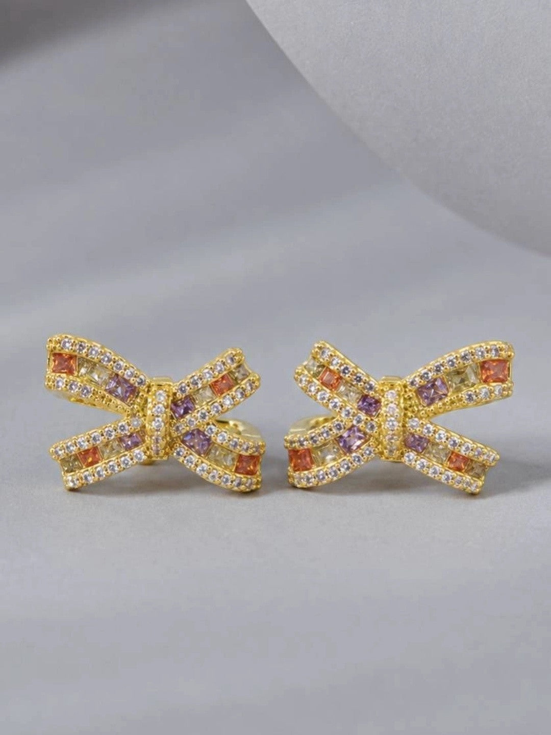 Pair of gold-plated bow stud earrings with multicolour zirconia, front view on grey surface