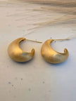 Pair of matte gold chunky crescent hoop earrings laid flat on a white marble surface with dried grass in background