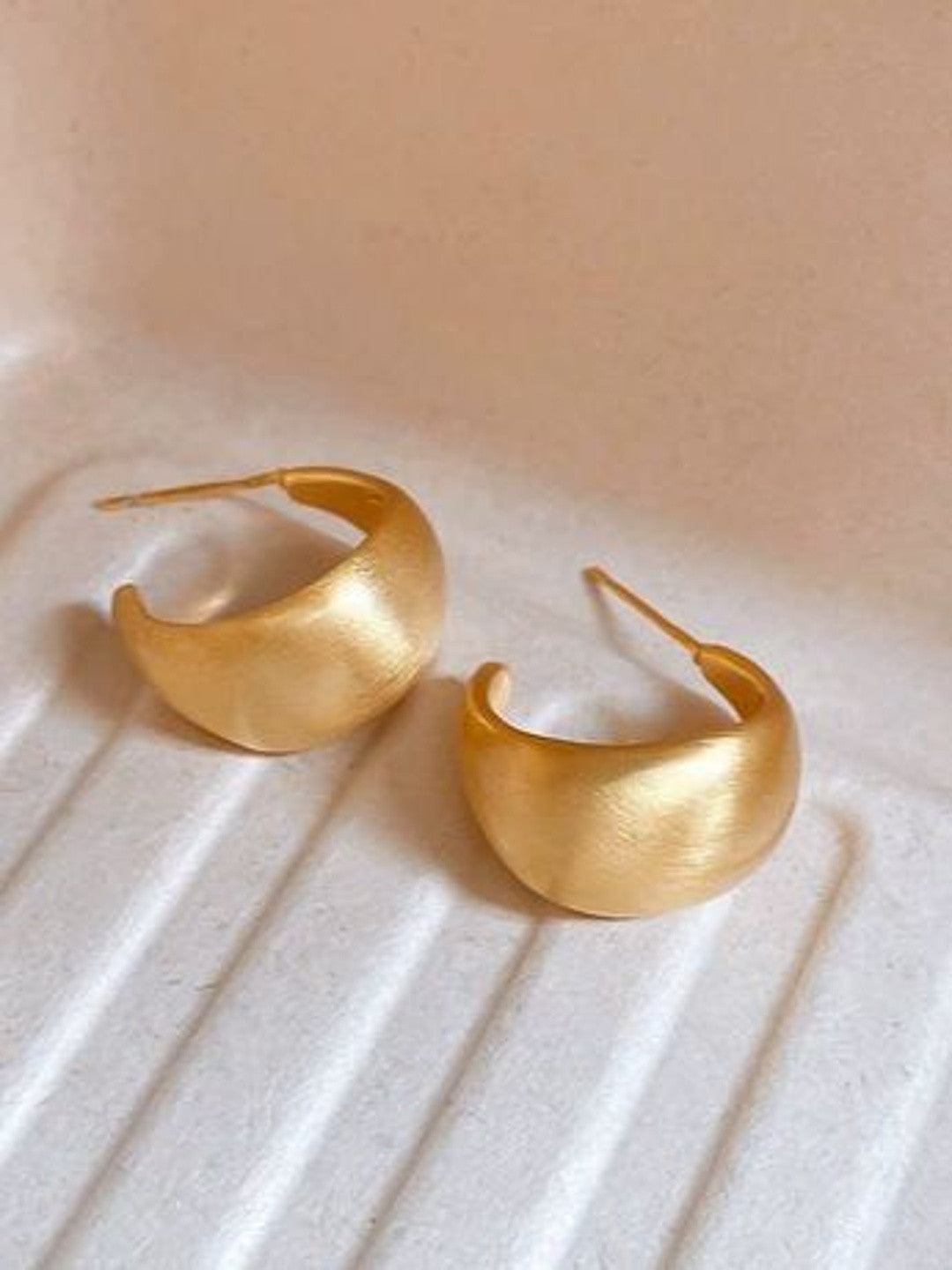 Two matte gold crescent hoop earrings placed side by side on a ridged white ceramic surface showing satin finish