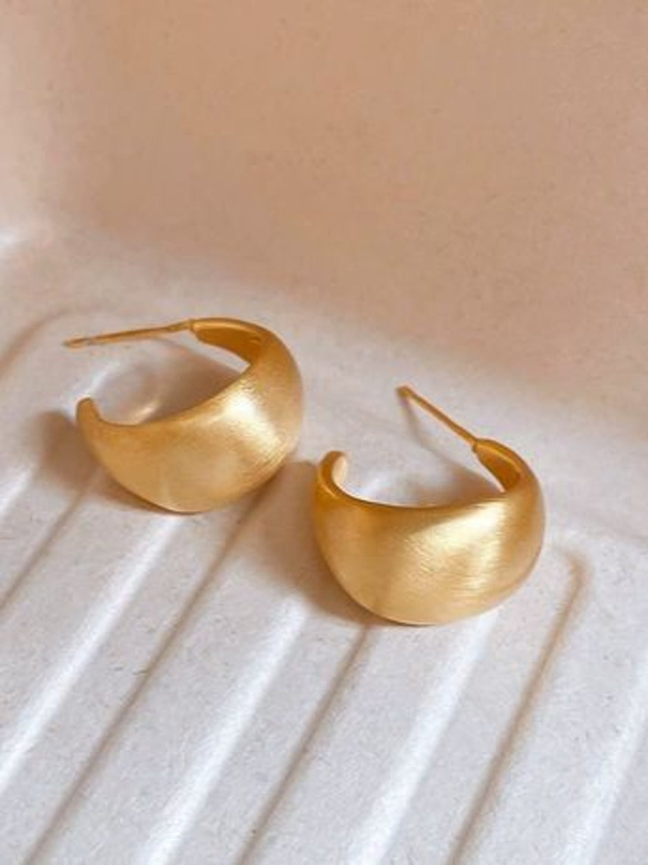 Two matte gold crescent hoop earrings placed side by side on a ridged white ceramic surface showing satin finish