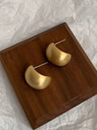 Pair of matte gold chunky hoop earrings resting on a dark walnut wooden tray against crinkled white fabric