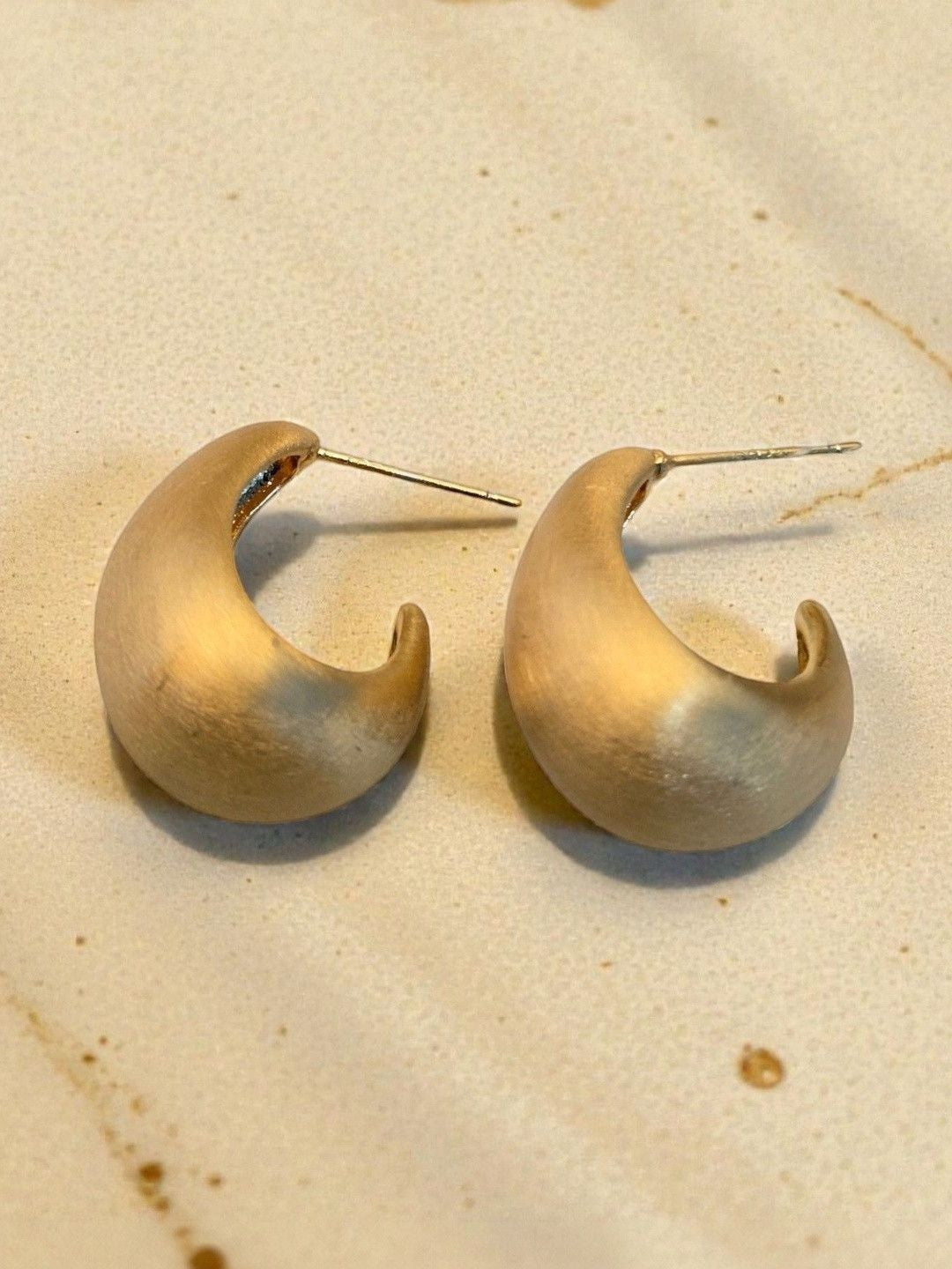 Two matte gold crescent hoop earrings with post backs displayed on a cream textured stone surface in natural light