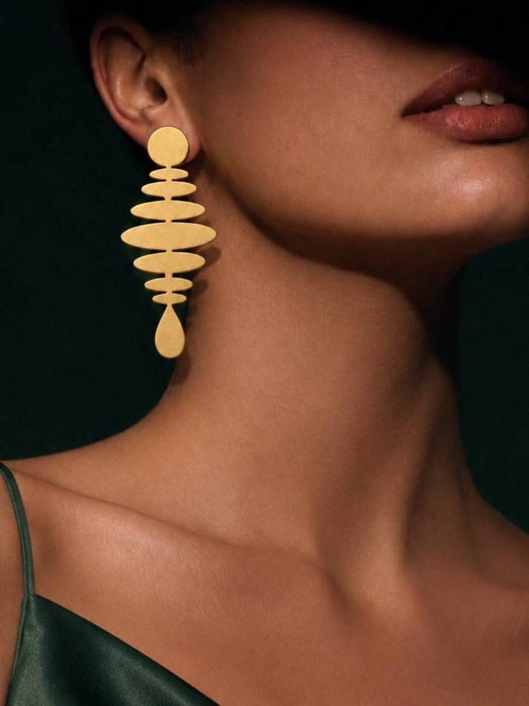 Model wearing one matte gold geometric drop earring against dark background with green silk top