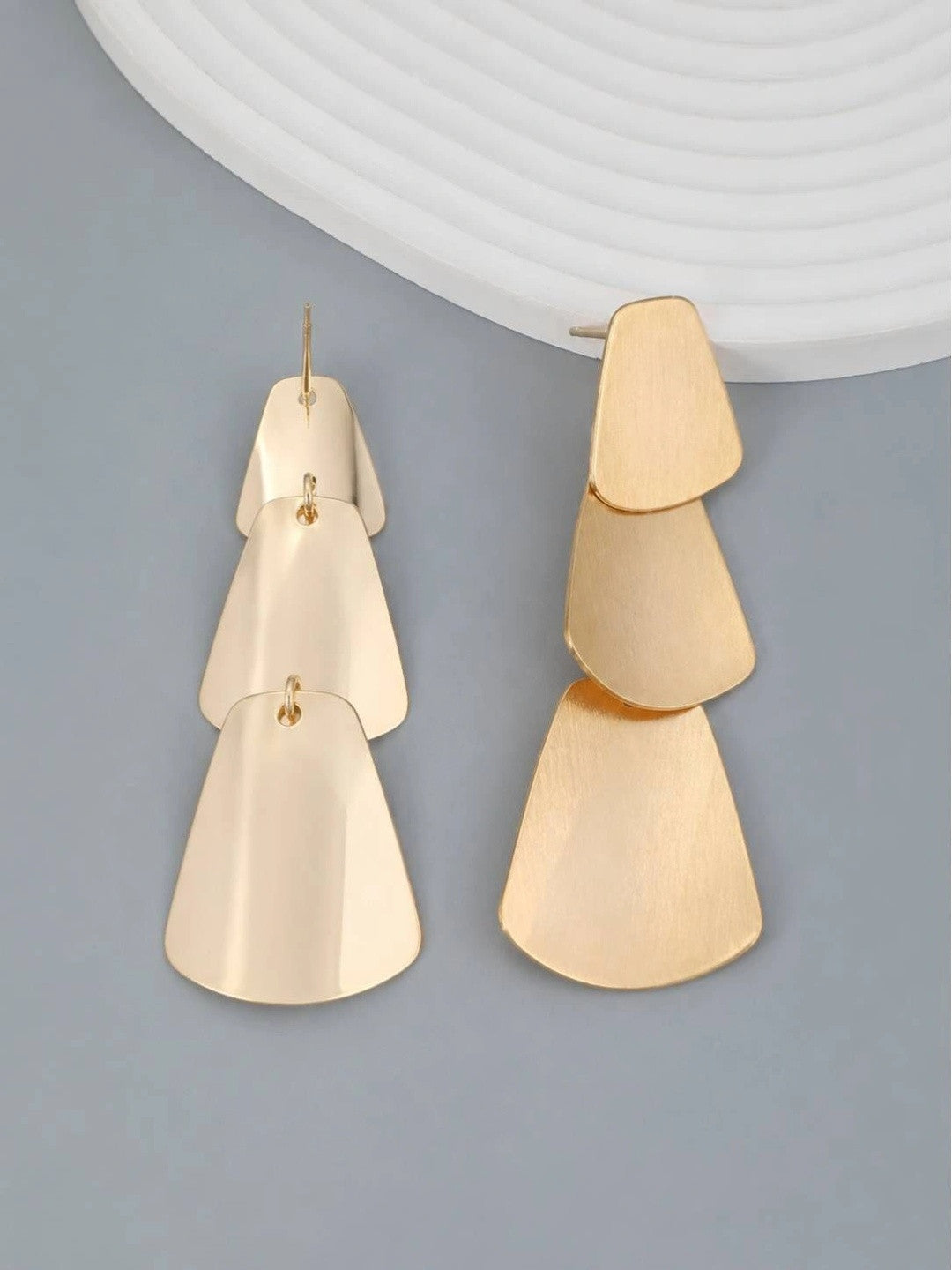 Pair of gold-toned tiered geometric drop earrings shown flat on grey surface, one face-up and one at an angle
