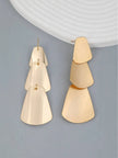 Pair of gold-toned tiered geometric drop earrings shown flat on grey surface, one face-up and one at an angle