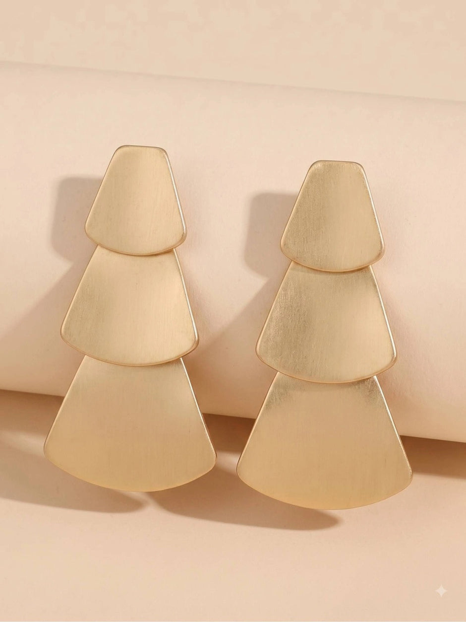 Two brushed gold geometric tiered drop earrings leaning against a beige cylindrical prop