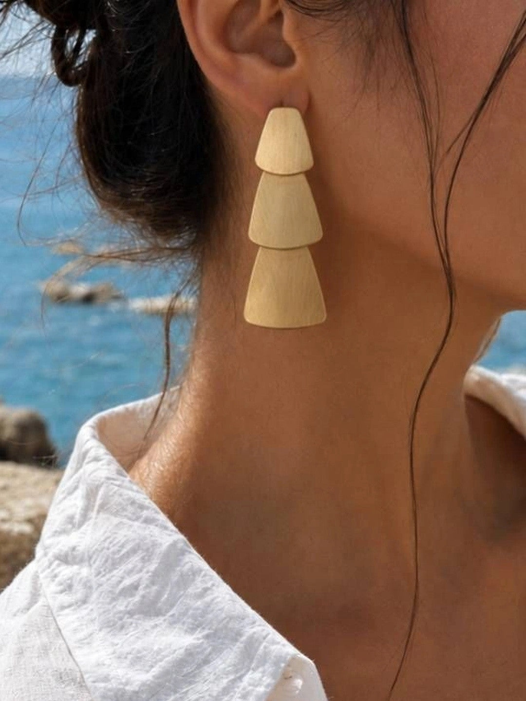 Model wearing gold matte tiered geometric drop earring on left ear with white off-shoulder top, ocean in background