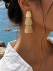 Model wearing gold matte tiered geometric drop earring on left ear with white off-shoulder top, ocean in background