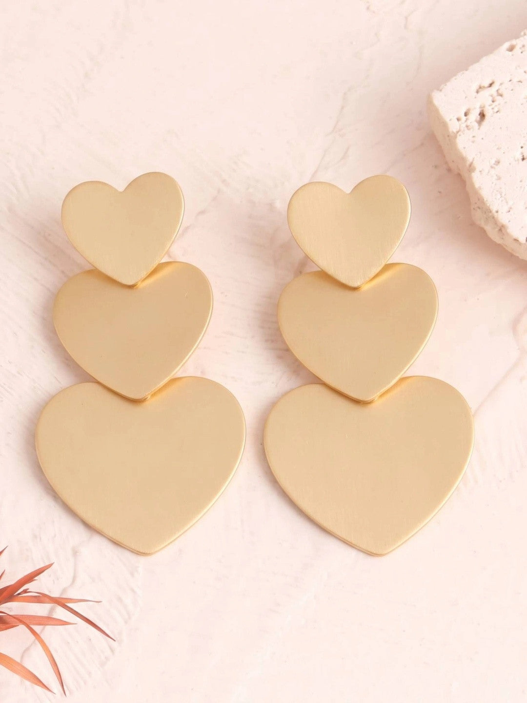 Pair of matte gold triple heart drop earrings laid flat on a pink marble surface