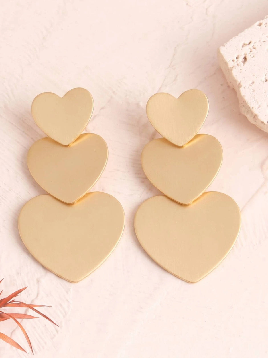 Pair of matte gold triple heart drop earrings laid flat on a pink marble surface