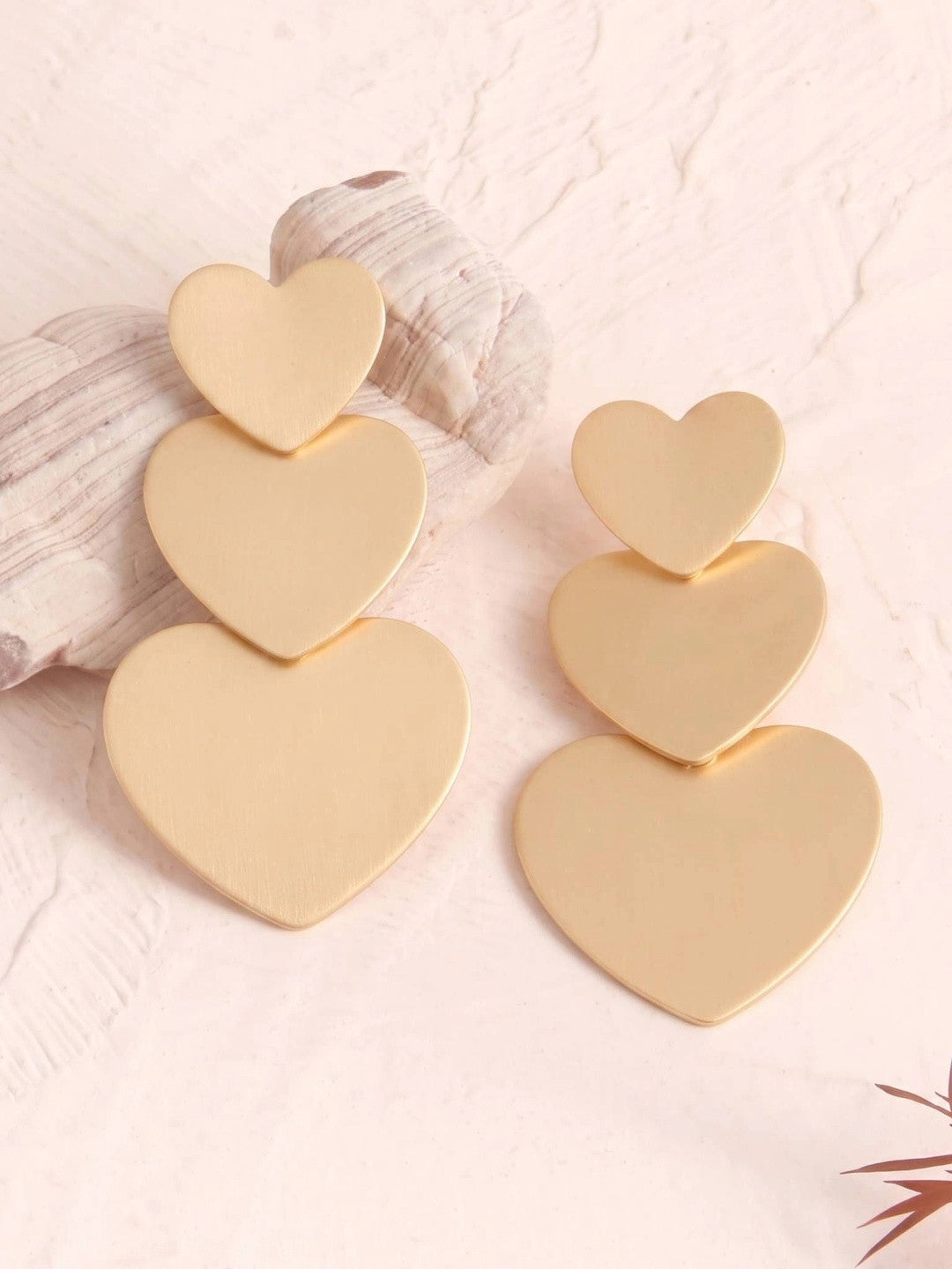 Two matte gold triple heart drop earrings displayed against a white stone and pink background