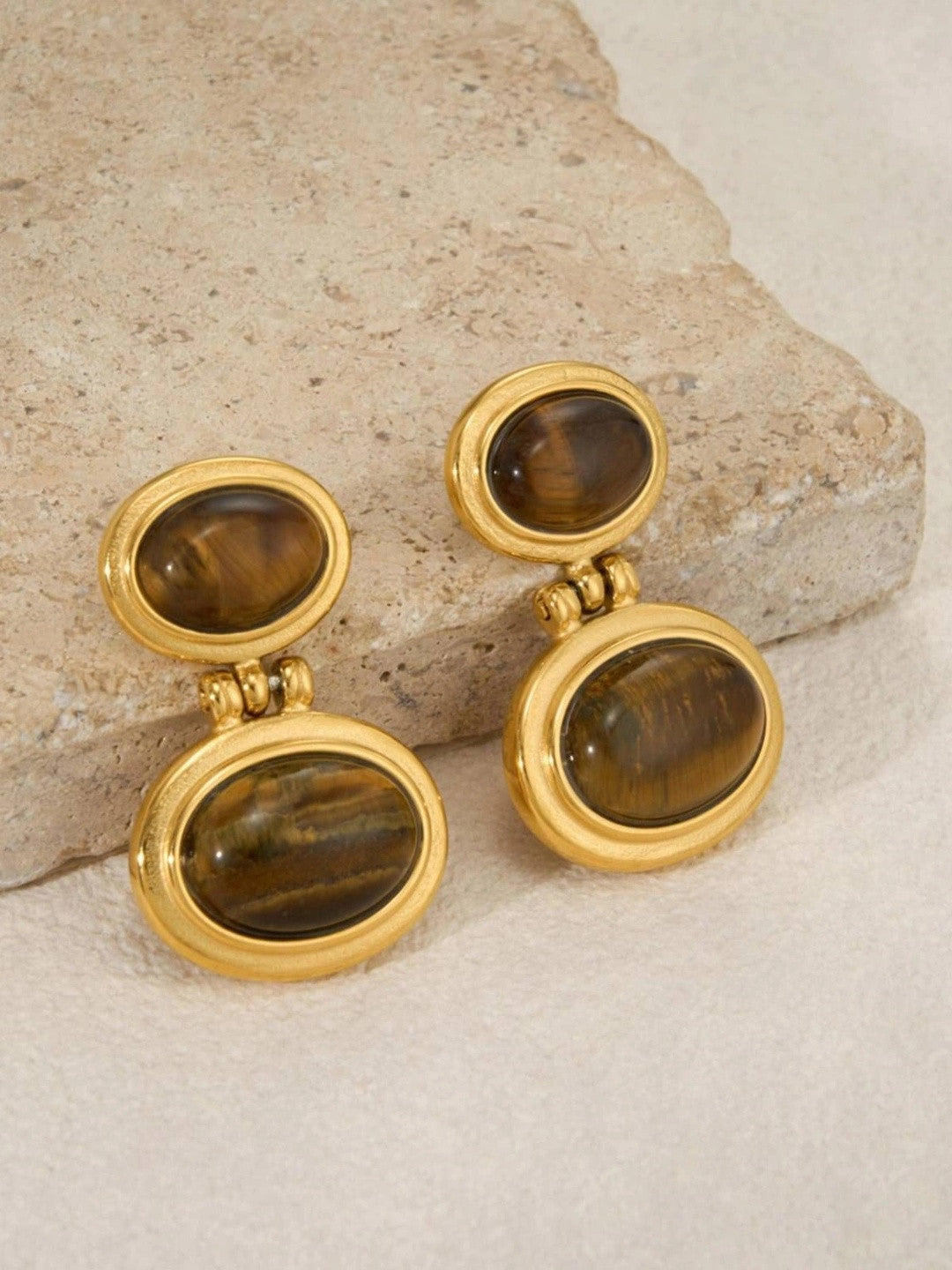 Pair of gold-plated double oval drop earrings with tiger eye stones resting on a stone surface
