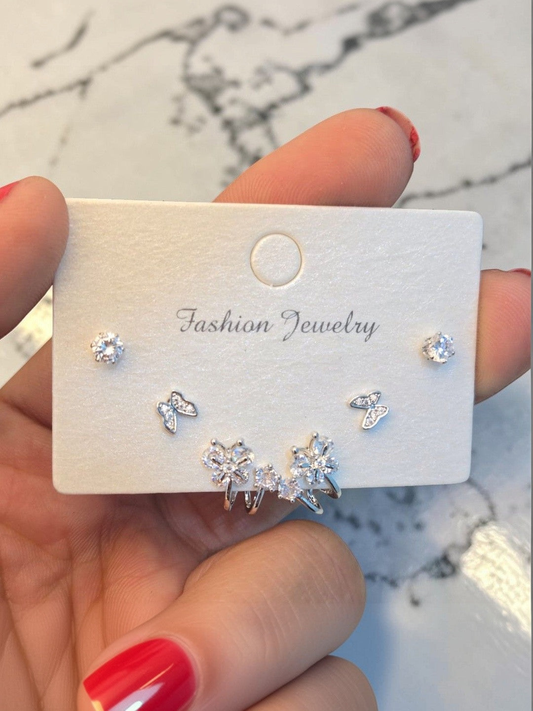 Hand holding a white jewellery card displaying three pairs of silver CZ earrings against a marble background