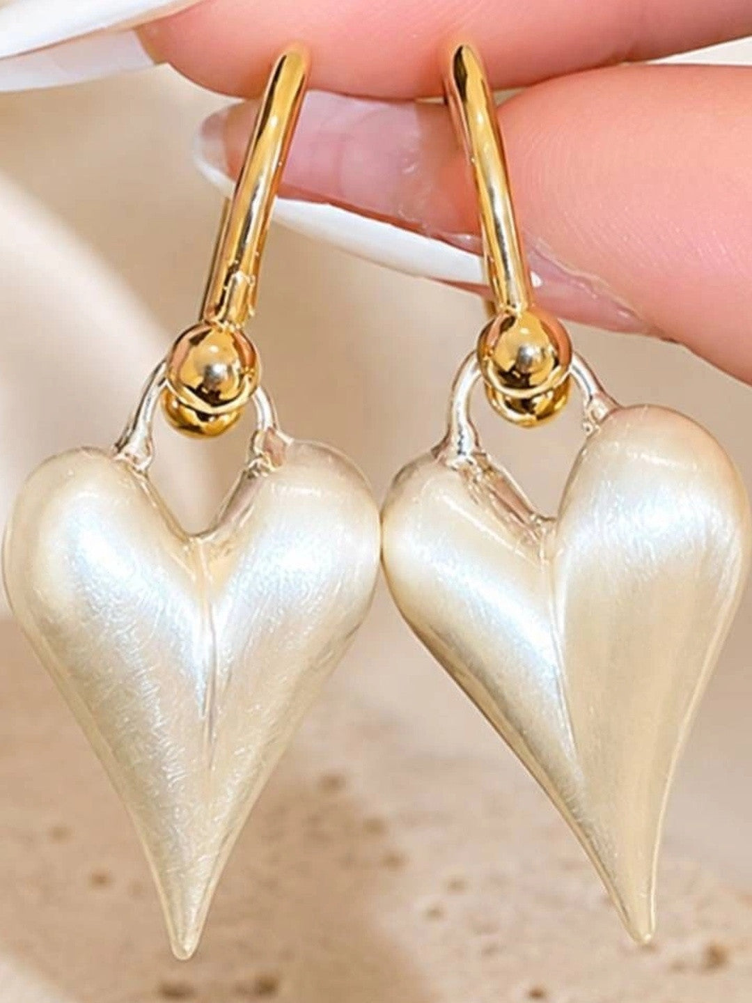 Two pearl-lustre puffy heart drop earrings held between fingers against a soft cream background