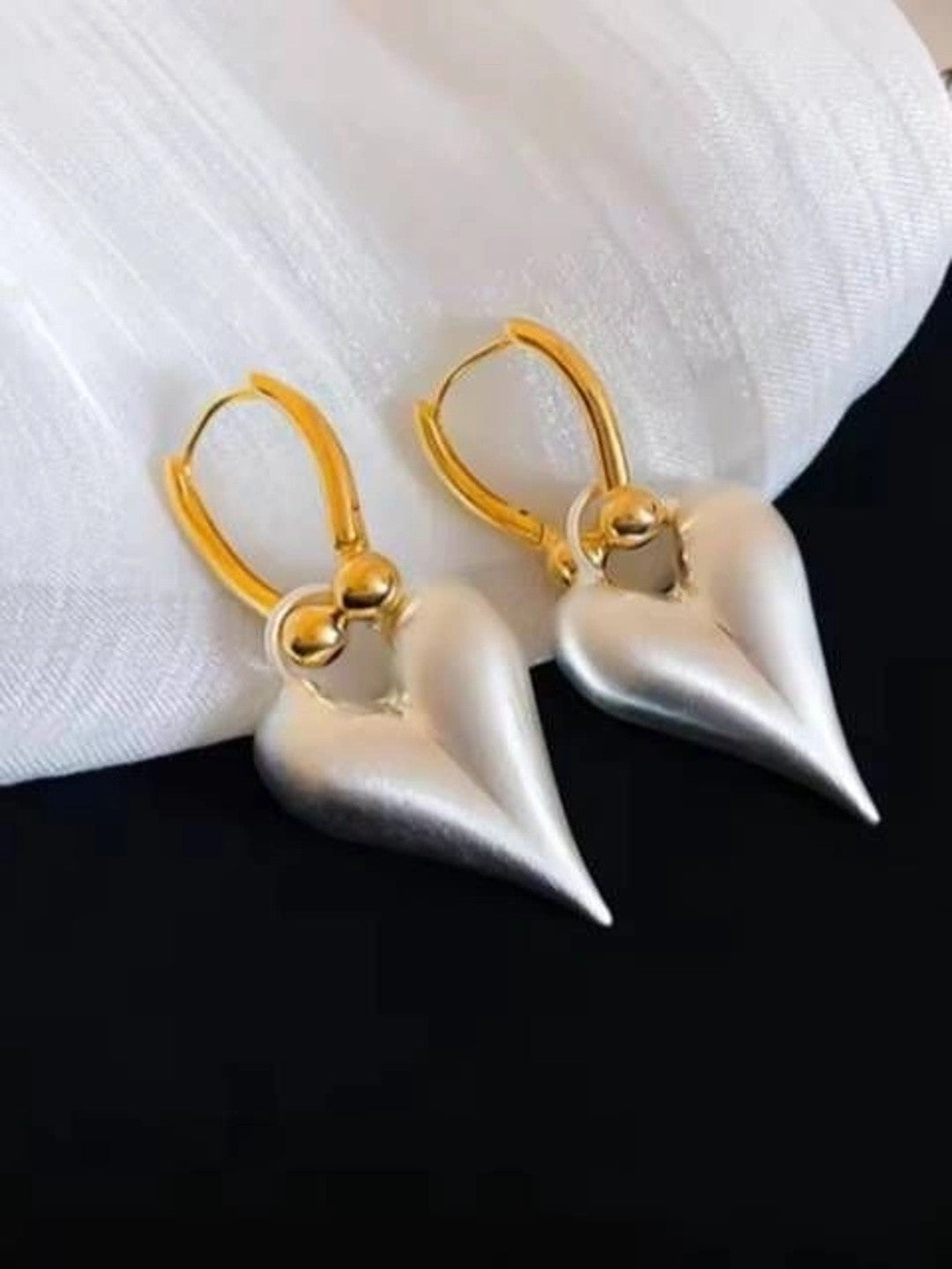 Pair of silver-tone matte heart drop earrings with gold lever-back hoops on a white fabric surface