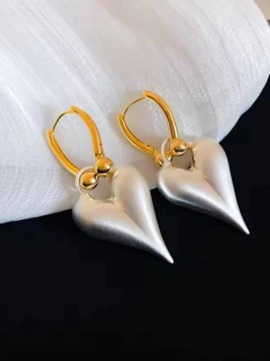 Pair of silver-tone matte heart drop earrings with gold lever-back hoops on a white fabric surface
