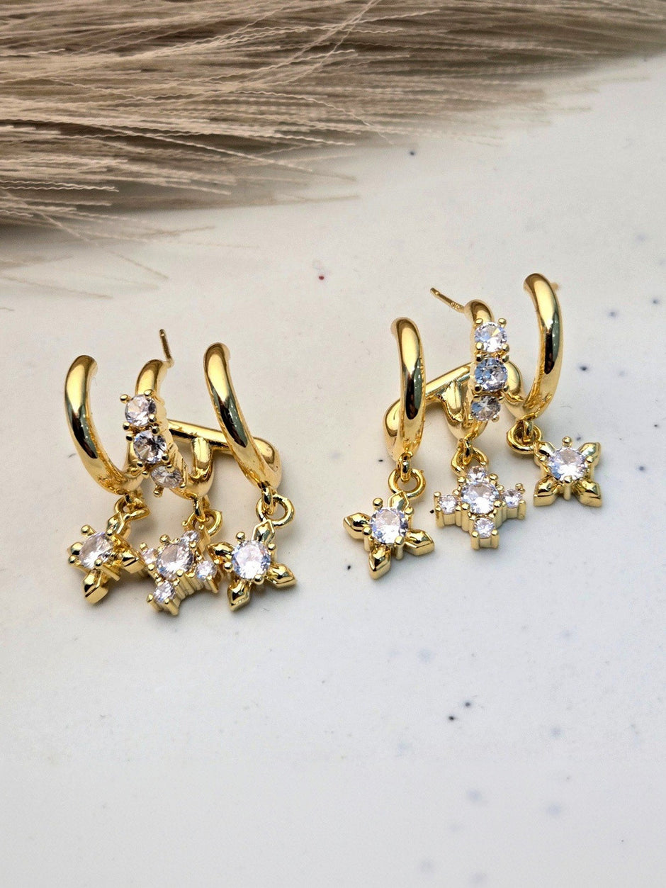Two pairs of gold-plated crystal star drop hoop earrings displayed on a beige stone surface