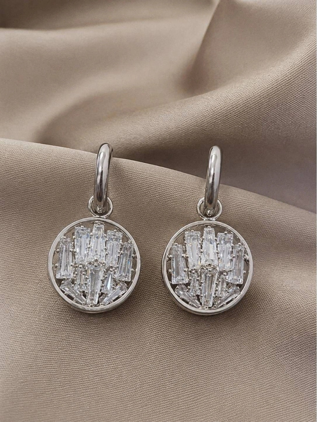Pair of silver circular drop earrings with baguette crystal clusters on a beige fabric background