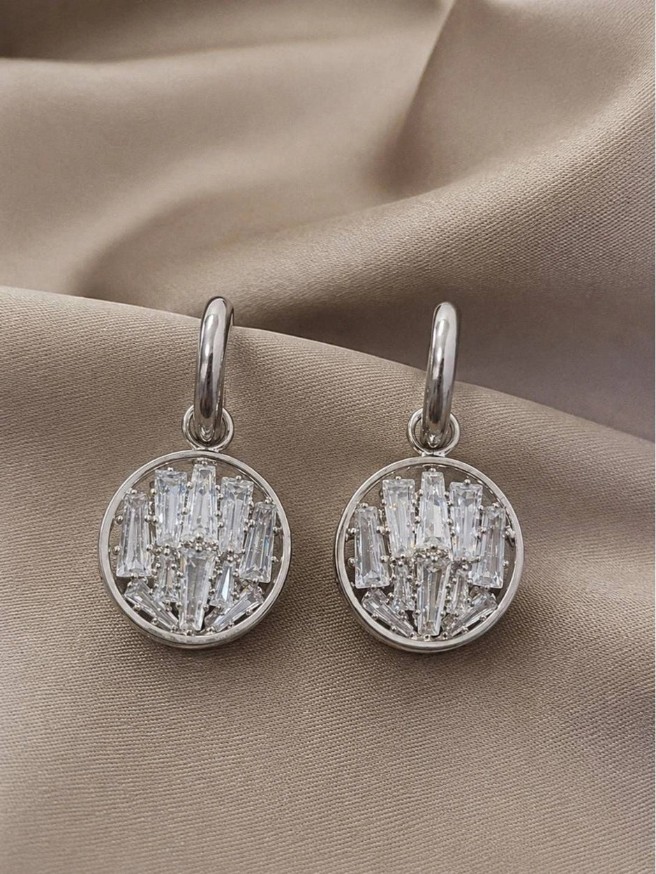 Pair of silver circular drop earrings with baguette crystal clusters on a beige fabric background