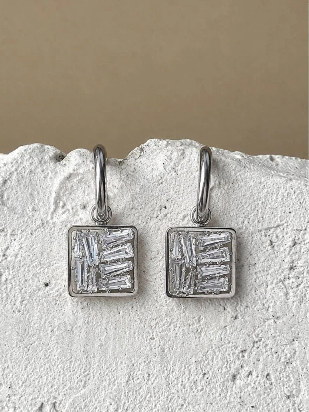 Pair of silver-finish square drop earrings with baguette zirconia stones displayed on a white stone surface