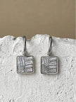 Pair of silver-finish square drop earrings with baguette zirconia stones displayed on a white stone surface