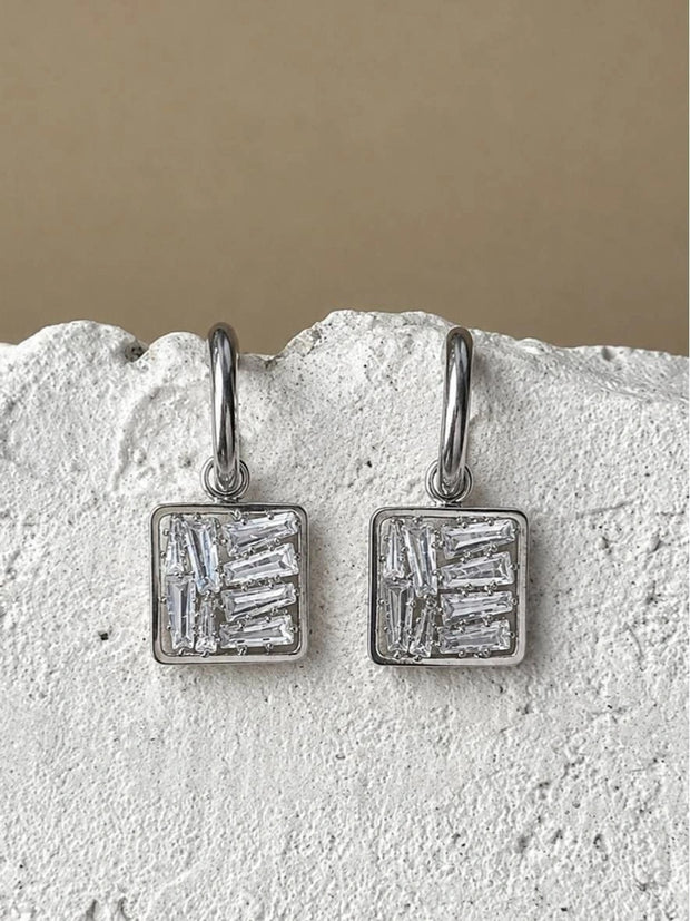 Pair of silver-finish square drop earrings with baguette zirconia stones displayed on a white stone surface